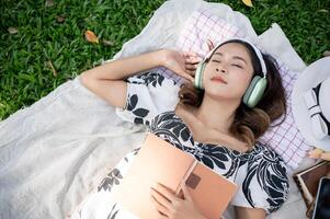 un asiático mujer toma un siesta mientras escuchando a música en su auriculares y leyendo un libro en un parque. foto