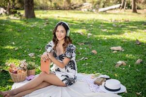 atractivo asiático mujer es escuchando a música en su auriculares y leyendo un libro mientras hacer un picnic. foto