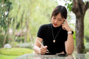 un hermoso, pensativo asiático mujer se sienta a un al aire libre mesa en un jardín utilizando su digital tableta. foto