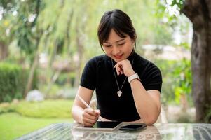 un atractivo asiático mujer se sienta a un al aire libre mesa en un jardín o patio interior utilizando su digital tableta. foto