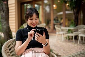 A cheerful, happy Asian woman sits at an outdoors table in the garden of a cafe using her smartphone photo