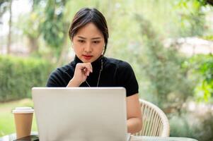 un hermoso, grave asiático mujer se sienta a un mesa en un jardín utilizando su computadora portátil, planificación su trabajar. foto