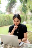 An attractive Asian woman sits at a table in a garden working, reading online document on her laptop photo