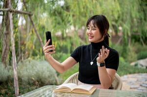 A cheerful Asian woman is talking on a call on her smartphone while relaxing in a garden. photo