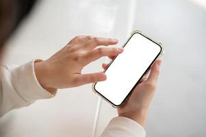 A woman in a white sweater holding a smartphone white-screen mockup over a white table indoors. photo