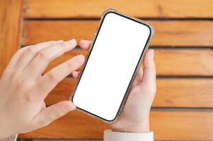 Top view image of a woman's hand holding a white-screen smartphone mockup over a wooden table. photo