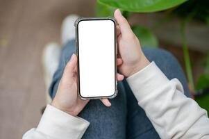 A close-up image of a woman is sitting next to a houseplant indoors and using her smartphone. photo