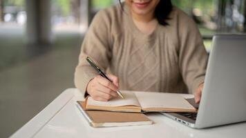 A cropped image of an Asian woman taking notes in her notebook while working on her laptop computer. photo