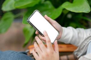 A close-up image of a woman is sitting next to a houseplant indoors and using her smartphone. photo