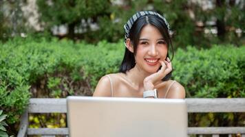 An attractive Asian woman smiles at the camera, sits at a table in a garden with her laptop computer photo