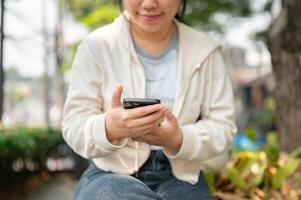 A cropped shot of a young Asian woman sits on a bench in a city park and uses her smartphone. photo