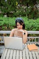 un dudoso asiático mujer trabajando en su ordenador portátil en un jardín, luchando con un bajo Internet conexión. foto