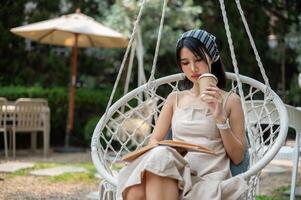 A woman is sipping coffee and focusing on reading a book while chilling on a swing in the garden. photo