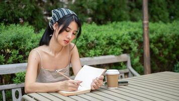 A beautiful Asian woman in a cute dress focuses on writing in her diary in the green garden. photo