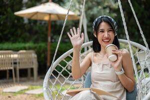 An attractive Asian woman chilling on a swing in a garden, waving her hand and smiling at the camera photo