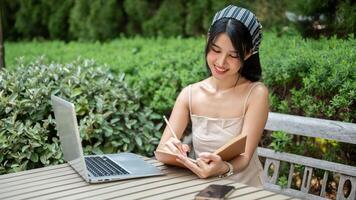 un mujer trabajando remotamente en un jardín, tomando notas en su libro y trabajando en su ordenador portátil computadora. foto