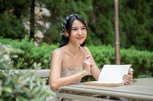 un hermosa asiático mujer en un linda vestir es soñador mientras escritura su diario en el verde jardín. foto