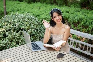 A woman waves her hand and smiles at camera while sitting at a table with her laptop in the garden. photo