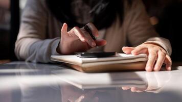 A cropped image of a woman holding a pen and using her smartphone at a table. photo
