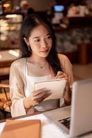 A focused female college student sits in a coffee shop taking notes, studying online on her laptop. photo