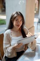 A beautiful Asian woman holding a digital tablet and smiling at the camera at a cafe. photo