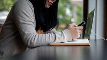 un recortado lado ver imagen de un asiático mujer utilizando su teléfono inteligente mientras sentado en un café tienda. foto