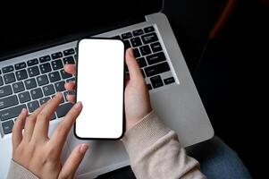 Top view image of a woman's hand holding a white-screen smartphone mockup over an opened laptop. photo