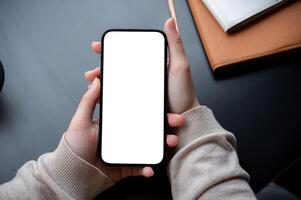 Top view image of a woman's hand holding a white-screen smartphone mockup over a modern office desk. photo