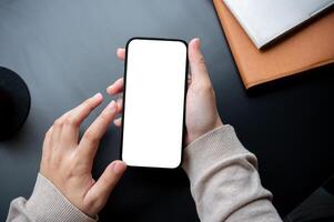 Top view image of a woman holding a smartphone white-screen mockup over an office desk workspace. photo