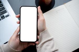 Top view image of a woman's hand holding a white-screen smartphone mockup over a modern office desk. photo