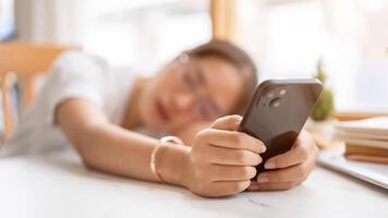 A tired, bored Asian woman leaning on the table and holding her smartphone, waiting for the messages photo