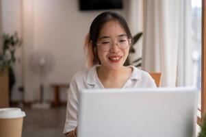 un joven asiático mujer es hablando, teniendo un en línea reunión mientras trabajando remotamente a un café tienda. foto