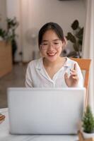 un joven asiático mujer es hablando, teniendo un en línea reunión mientras trabajando remotamente a un café tienda. foto