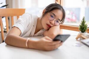 A tired, bored Asian woman leaning on the table and holding her smartphone, waiting for the messages photo