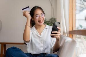 A cheerful Asian woman holding a credit card and a smartphone, enjoying shopping online at home. photo