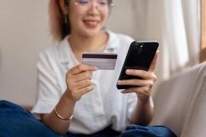 A young, cheerful Asian woman sits on a sofa, using a mobile banking app, shopping online at home. photo