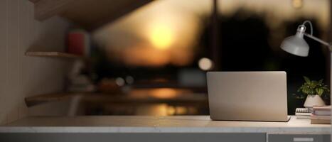 A laptop computer on a table in a room with dim lights from a lamp and sunset through the window. photo