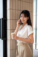 un atractivo asiático mujer participación su café taza mientras hablando en el teléfono en el edificio. foto