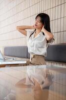 An overworked Asian businesswoman is feeling tired, massaging her neck while working on her laptop. photo