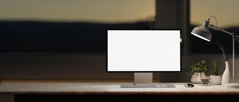 A computer desk in a modern, dark room in the evening features a white-screen computer mockup. photo