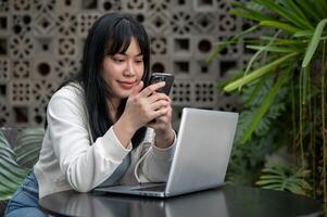 A young happy Asian woman is using her smartphone while working remotely at a cafe. photo