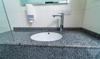 Public clinic bathroom with soap dispenser on tiled wall with mirror. Granite sink for washing hands and face. Closeup. photo