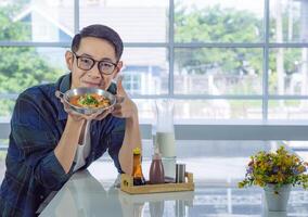 Young man wearing glasses holding the fried egg served on a pan with colorful toppings served on a pan while sitting on a chair. photo