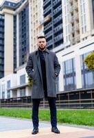 Man in gray coat and black shirt on background of business skyscrapers, looking at the camera. Businessman in coat standing near international business centre. photo