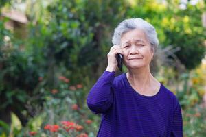 Portrait of a senior woman with short gray hair using a smartphone while standing in a garden. Space for text. Concept of aged people and communication photo