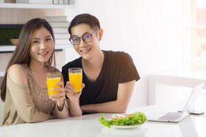 asiático joven Pareja participación un vaso de Fresco naranja jugo para salud con vegetal ensalada, computadora portátil, y un teléfono inteligente metido en el mesa mientras sentado en el cocina. concepto de amor Pareja foto