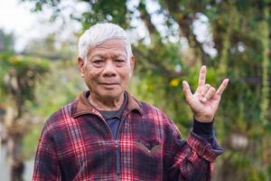 Portrait of a senior man with short gray hair showing fingers I love you a symbol, smliling, and looking at the camera while standing outdoors. Concept of aged people and healthcare photo