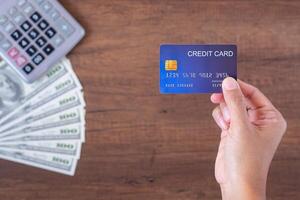 Close-up of hand holding a blue credit card with US banknotes and a calculator background placed on a wooden table. Space for text. Wood texture background. Business and payment concept photo