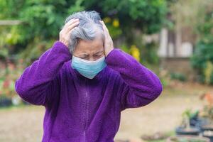 mayor asiático mujer vistiendo un cara máscara y dolor de cabeza mientras en pie en un jardín. máscara para proteccion virus, COVID-19, coronavirus, bacterias, polvo. concepto de Envejecido personas y cuidado de la salud foto