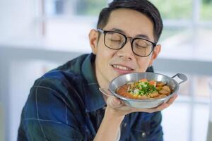 Young man wearing glasses holding the fried egg served on a pan with colorful toppings served on a pan while sitting on a chair. photo